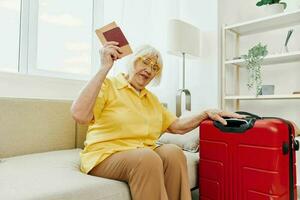 contento anziano donna con passaporto e viaggio biglietto confezionato un' rosso valigia, vacanza e Salute cura. sorridente vecchio donna con gioia seduta su il divano prima il viaggio sollevato sua mani su nel la gioia. foto