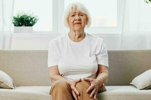 anziano donna acuto dolore nel sua gamba seduta su il divano, Salute i problemi nel vecchio età, povero qualità di vita. nonna con grigio capelli detiene su per sua dolorante ginocchio, i problemi con articolazioni e legamenti. foto