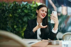 elegante moda donna blogger turista si siede nel un' bar con un' Telefono nel sua mani legge un' Messaggio, mobile comunicazione e Internet su un' viaggio, video chiamata, libero professionista opera in linea, Sorridi con denti foto