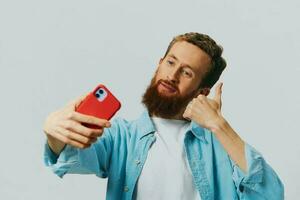 uomo fricchettone con Telefono nel mano blogger Sorridi su grigio sfondo nel blu camicia e bianca maglietta, parlando su il Telefono e selfie foto