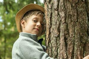 un' contento caucasico ragazzo nel un' cappello abbracci un' albero. all'aperto passeggiate foto