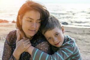 carino contento ragazzo abbracci il suo mamma di il mare. guardare in il telecamera foto