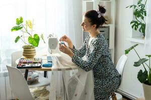 un' donna cuce tulle su un elettrico cucire macchina nel un' bianca moderno interno di un' Casa con grande finestre, Casa impianti. comfort nel il Casa, un' della casalinga passatempo foto