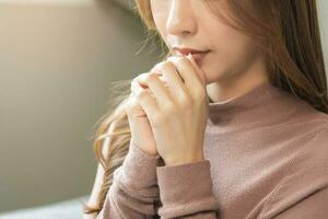 pieno di speranza rendere desiderio, calma asiatico giovane donna credenza, mano nel preghiera, seduta Meditare su divano a casa, cristiano ragazza preghiere per Dio per Chiedi, richiesta con chiuso occhi sognare per aiuto, grazie voi Dio. foto