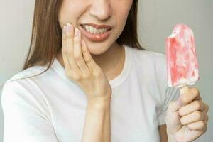 viso espressione sofferenza a partire dal sensibile denti e freddo, asiatico giovane donna, ragazza mano toccante sua guancia, sensazione male, dolore mangiare ghiaccio crema, lecca lecca. mal di denti molare dente a casa, dentale problema. foto