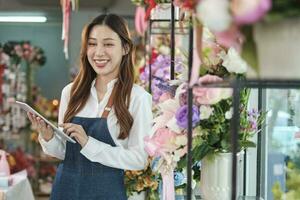 ritratto di uno bellissimo giovane asiatico femmina fioraio lavoratore nel grembiule con tavoletta nel luminosa e bellissimo fiore negozio negozio, in linea e-commerce opera, piccolo attività commerciale occupazione, contento pmi imprenditore. foto