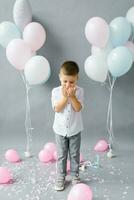 un' poco ragazzo-bambino colpi su coriandoli su un' grigio sfondo con colorato palloncini. vacanza. contento eccitato bambino su sua compleanno. foto