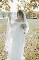 elegante bellissimo sposa sotto un' velo sta nel davanti e sembra a un' mazzo nel un' bianca nozze vestire. il sposa nel il bellissimo leggero sotto il albero. foto