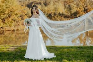 ritratto di un' bellissimo giovane sposa nel un elegante bianca vestito con un' lungo velo e un' mazzo di gypsophila vicino il lago nel morbido luce. foto