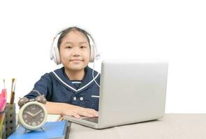 poco asiatico ragazza indossare cuffie e fare compiti a casa con il computer portatile in linea isolato foto