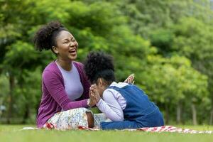 africano americano madre è ridendo mentre giocando polpetta torta con sua giovane figlia mentre avendo un' estate picnic nel il pubblico parco per benessere e felicità concetto foto