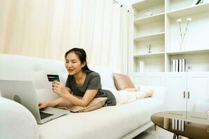donna asiatica in possesso di una carta di credito per lo shopping online mentre è seduta sul divano mentre è in vacanza a casa. foto