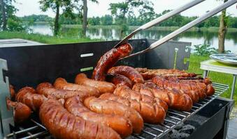 uomo svolta il salsiccia su il bbq con cucina pinza. persona grigliate nel il giardino. barbeque con salsicce e fritte pesce su il griglia. foto