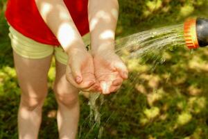 un' bambino mette il suo mani sotto acqua fluente a partire dal un' irrigazione tubo flessibile su un' caldo estate giorno foto