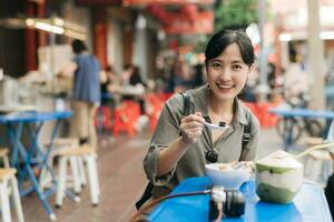 contento giovane asiatico donna zaino viaggiatore godendo strada cibo a Cina cittadina strada cibo mercato nel bangkok, Tailandia. viaggiatore controllo su lato strade. foto