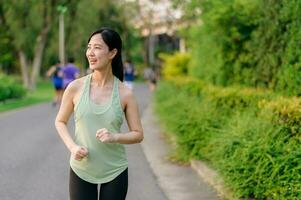 in forma asiatico giovane donna jogging nel parco sorridente contento in esecuzione e godendo un' salutare all'aperto stile di vita. femmina pareggiatore. fitness corridore ragazza nel pubblico parco. salutare stile di vita e benessere essere concetto foto