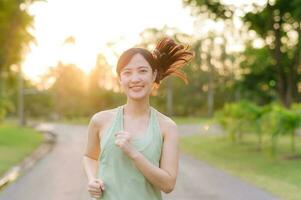 in forma asiatico giovane donna jogging nel parco sorridente contento in esecuzione e godendo un' salutare all'aperto stile di vita. femmina pareggiatore. fitness corridore ragazza nel pubblico parco. salutare stile di vita e benessere essere concetto foto