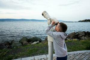 ragazzo guardare attraverso un' telescopio su il riva del mare a tramonto. foto