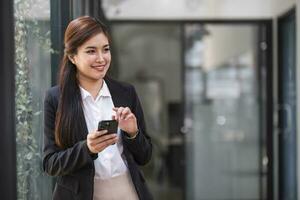 affascinante donna asiatica con un sorriso in piedi in possesso di documenti e telefono cellulare in ufficio. foto