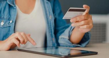 giovane donna asiatica che utilizza il tablet per lo shopping online e paga le bollette con la carta di credito nell'interno del soggiorno