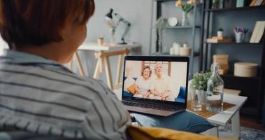 giovane ragazza asiatica che utilizza la videochiamata del laptop riunione parla con la famiglia anziana papà e mamma mentre lavora da casa seduto sul divano in soggiorno foto