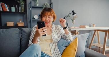 giovane signora asiatica che utilizza una videochiamata smart phone parla con la famiglia sul divano nel soggiorno di casa