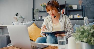 donna d'affari asiatica che utilizza laptop parla con i colleghi del piano in videochiamata mentre lavora da casa in soggiorno foto