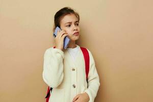 ritratto di contento sorridente bambino ragazza parlando su il Telefono con un' zaino beige sfondo foto