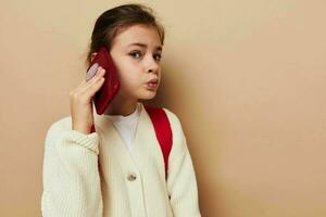 ritratto di contento sorridente bambino ragazza parlando su il Telefono con un' zaino beige sfondo foto