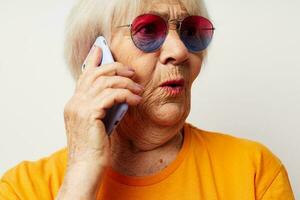 anziano donna nel casuale maglietta occhiali da sole parlando su il Telefono isolato sfondo foto