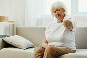 anziano donna acuto collo dolore seduta su il divano, Salute i problemi nel vecchio età, povero qualità di vita. foto