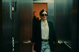 elegante donna nel nero giacca e occhiali da sole in posa nel ascensore, moda modello, buio cinematico leggero e colore, fascino Vintage ▾ foto