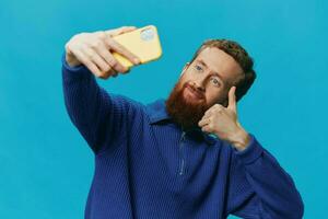 ritratto di un' uomo con un' Telefono nel il suo mani blogger prende selfie, su un' blu sfondo. comunicare in linea sociale media, stile di vita foto