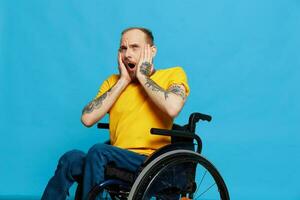 un' uomo nel un' sedia a rotelle sorpreso e Aperto bocca nel un' maglietta con tatuaggi su il suo braccia si siede su un' blu studio sfondo, un' pieno vita, un' vero persona foto