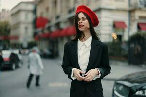 moda donna ritratto Sorridi con denti in piedi su il strada nel il città sfondo nel elegante Abiti rosso labbra e rosso berretto, viaggiare, cinematico colore, retrò Vintage ▾ stile, urbano moda stile di vita. foto