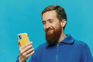 ritratto di un' uomo con un' Telefono nel il suo mani blogger prende selfie, su un' blu sfondo. comunicare in linea sociale media, stile di vita foto