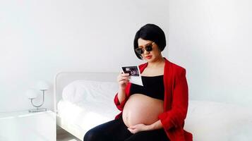 bellissimo e bella incinta donna indossare nero occhiali, seduta su letto, guardare a sua del bambino film immagine e toccante sua gonfiarsi. gravidanza 7-8 mesi, maternità, amore, aspettativa e cura. foto