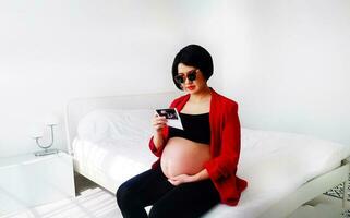 bellissimo e bella incinta donna indossare nero occhiali, seduta su letto, guardare a sua del bambino film immagine e toccante sua gonfiarsi. gravidanza 7-8 mesi, maternità, amore, aspettativa e cura. foto