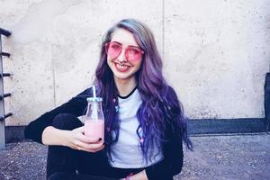felice bella adolescente con occhiali da sole rosa beve e gode di una bevanda rosa seduto su un terreno urbano foto