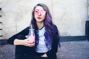 felice bella adolescente con occhiali da sole rosa beve e gode di una bevanda rosa seduto su un terreno urbano foto