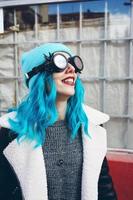 Ritratto di una giovane donna punk o gotica sorride con i capelli di colore blu e indossa occhiali steampunk neri e berretto di lana blu in una strada urbana all'aperto