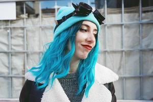 Ritratto di una giovane donna punk o gotica sorride con i capelli di colore blu e indossa occhiali steampunk neri e berretto di lana blu in una strada urbana all'aperto