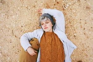 Un avventuriero giovane donna caucasica sdraiato su un terreno di sabbia accanto a uno zaino che indossa un maglione di lana e berretto con gli occhi chiusi e l'arancione come colore principale foto