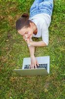 giovane donna dire bugie su verde erba prato nel città parco Lavorando su il computer portatile pc computer. libero professionista attività commerciale concetto foto