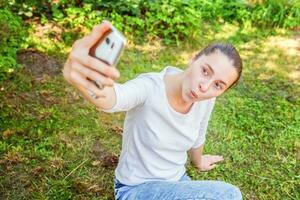 giovane ragazza prendere autoscatto a partire dal mani con Telefono mostrando divertente viso seduta su parco o giardino sfondo foto