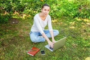 giovane donna seduta sul prato di erba verde nel parco cittadino che lavora su un computer portatile. concetto di business freelance foto