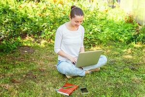 giovane donna seduta sul prato di erba verde nel parco cittadino che lavora su un computer portatile. concetto di business freelance foto