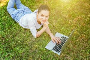 giovane donna dire bugie su verde erba prato nel città parco Lavorando su il computer portatile pc computer. libero professionista attività commerciale concetto foto