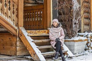 giovane donna vestita calorosamente in posa sotto il portico di una casa in legno nel villaggio. vacanze invernali in campagna