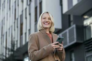 bellissima bellissimo giovane donna con bionda capelli messaggistica su il smart-phone a il città strada sfondo. bella ragazza avendo inteligente Telefono conversazione foto